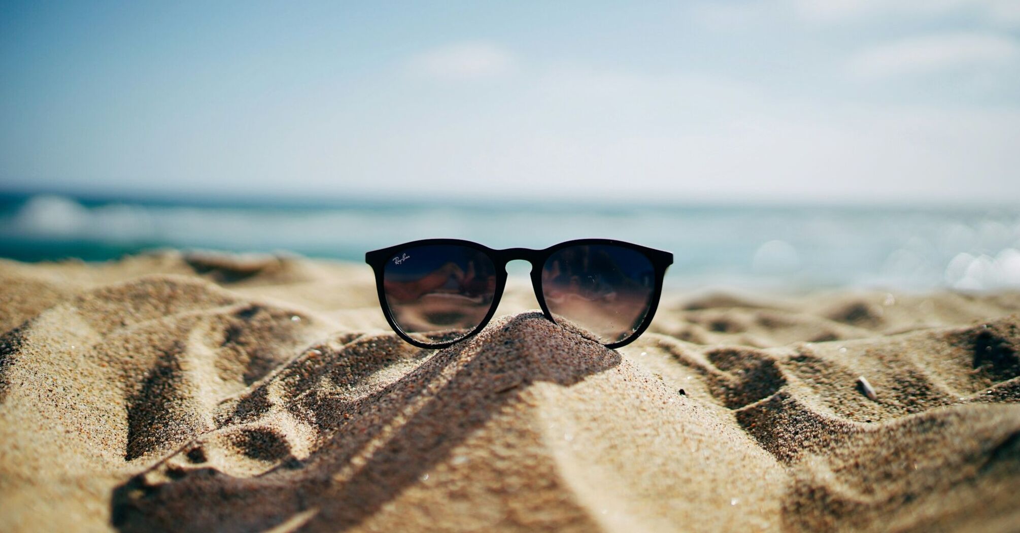 A pair of sunglasses lying on a sandy beach with a blurred ocean background