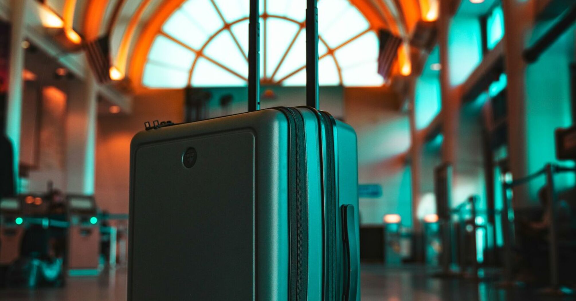 A sleek, teal suitcase stands in a modern airport under a curved, illuminated archway