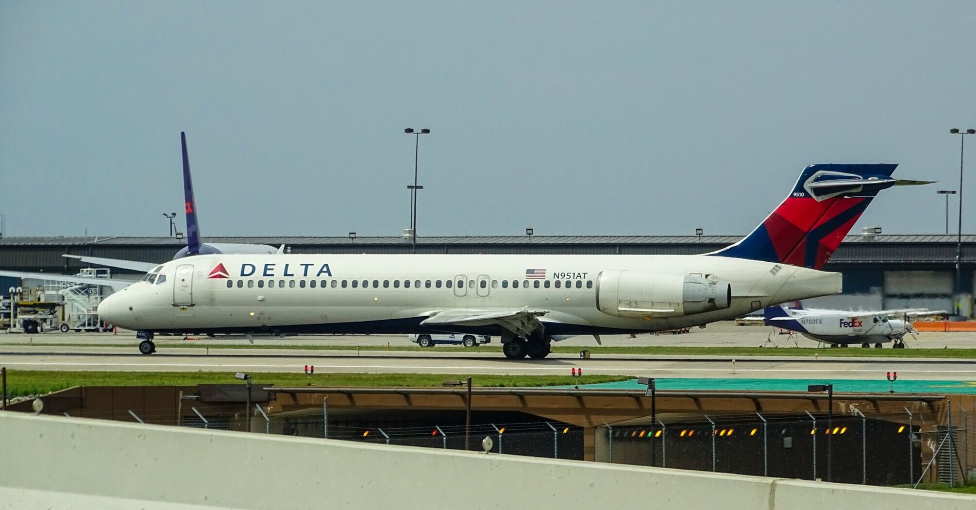 A Delta Airlines plane taxiing on the tarmac at an airport, with other aviation activity in the background