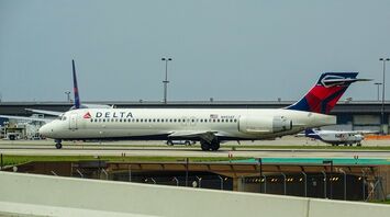 A Delta Airlines plane taxiing on the tarmac at an airport, with other aviation activity in the background