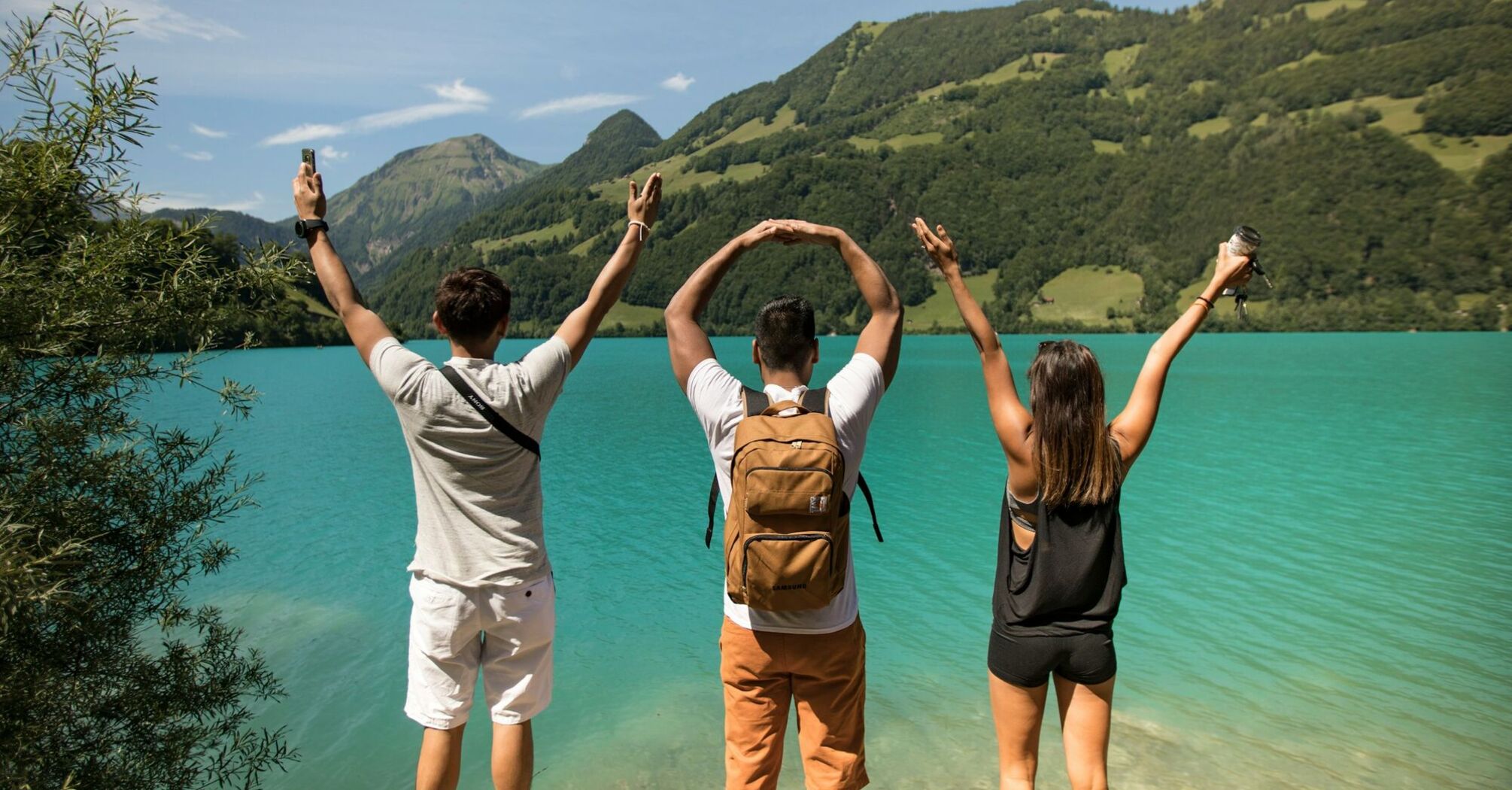 Three friends forming “WoW” with their hands while overlooking a turquoise lake surrounded by lush green hills