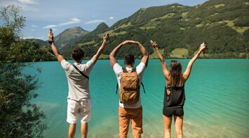 Three friends forming “WoW” with their hands while overlooking a turquoise lake surrounded by lush green hills