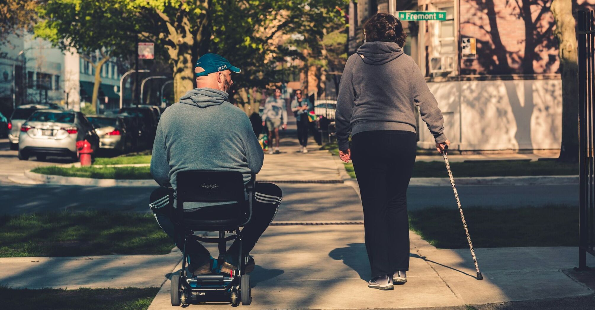 An individual in a motorized wheelchair and another person walking with a cane on a city sidewalk