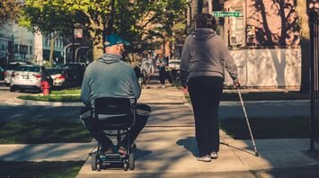 An individual in a motorized wheelchair and another person walking with a cane on a city sidewalk