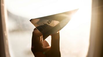 A hand holding a paper airplane up to an airplane window with sunlight streaming through
