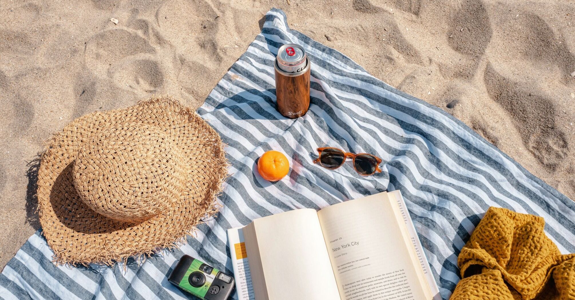 Beach setup with a hat, book, camera, sunglasses, and drink