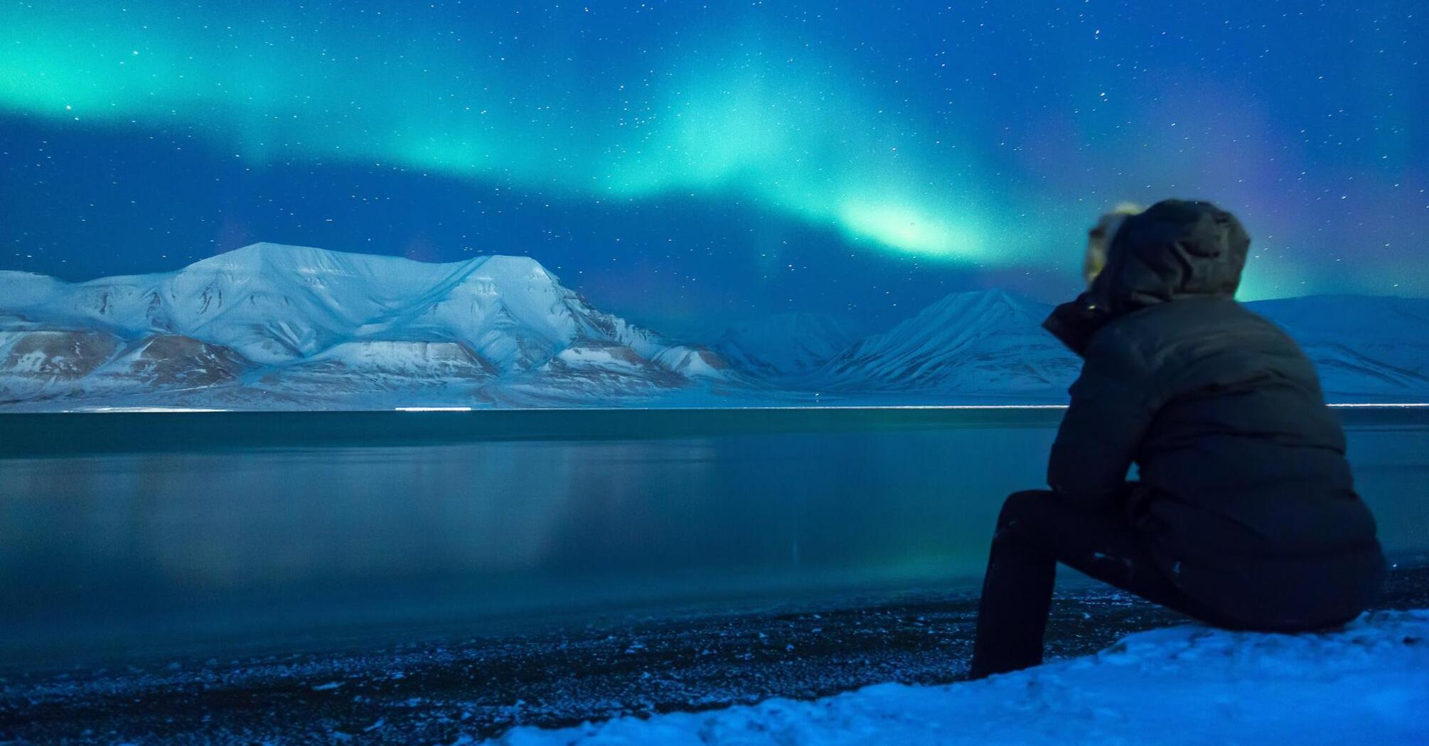 Man sits by a lake with the northern lights in the sky