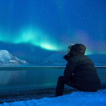 Man sits by a lake with the northern lights in the sky
