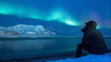 Man sits by a lake with the northern lights in the sky