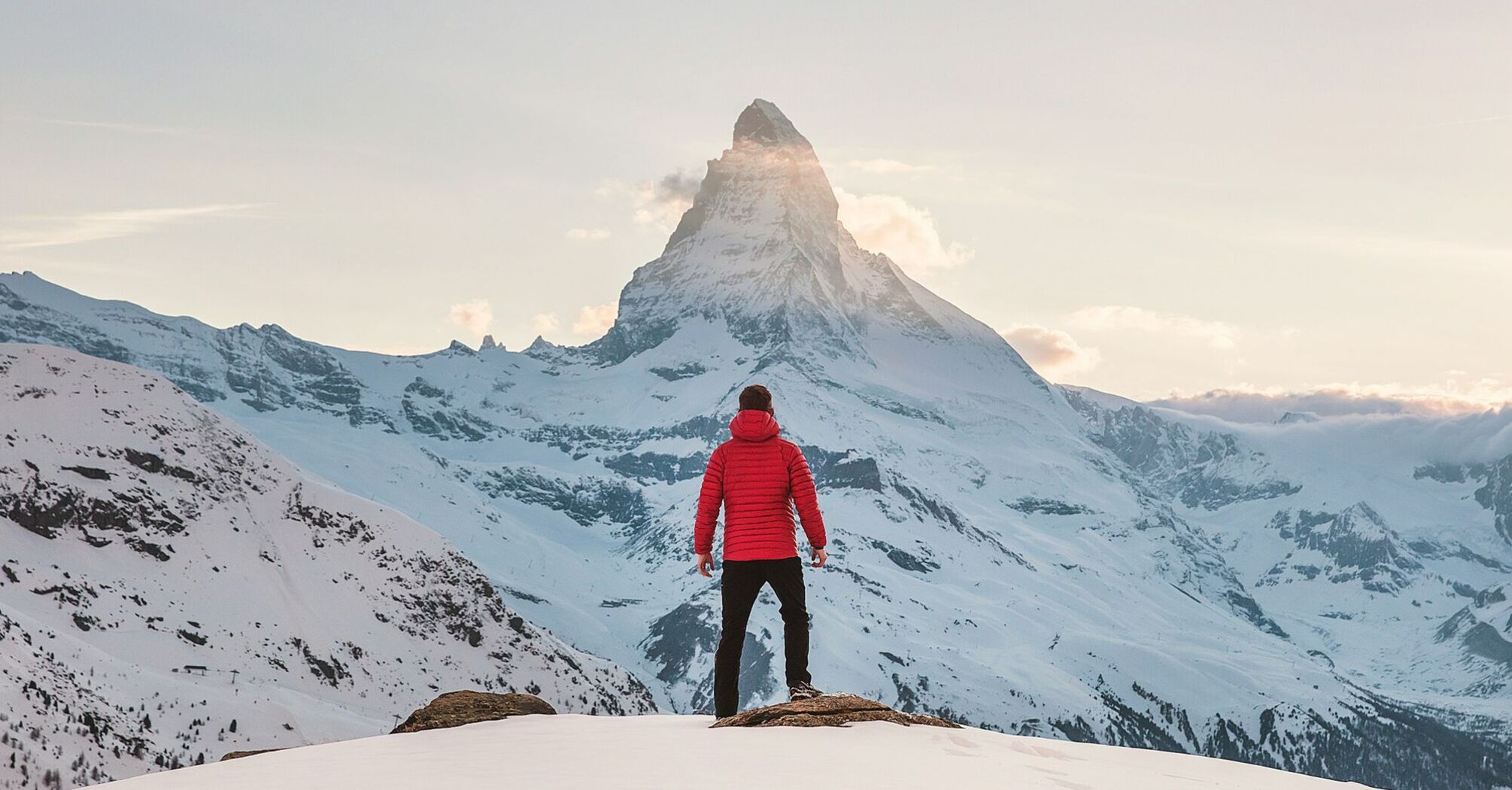 A person in a red jacket standing on a snowy mountain, gazing at a distant peak