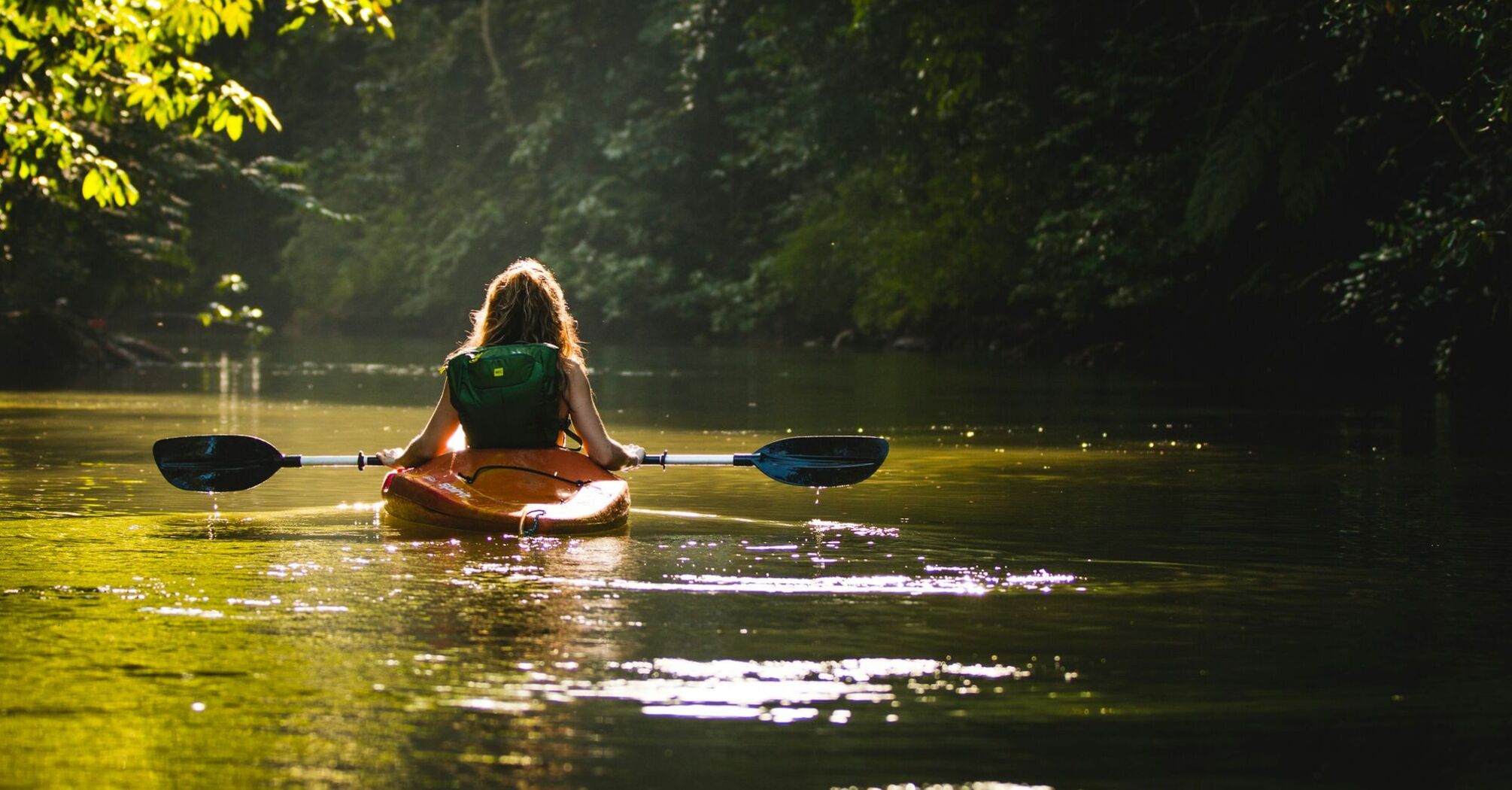 Woman kayaking through a serene, forested river with sunlight filtering through the trees