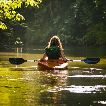 Woman kayaking through a serene, forested river with sunlight filtering through the trees