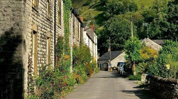 A picturesque street in a village with stone houses and a backdrop of green hills under a clear blue sky