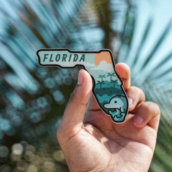 A person holding a colorful Florida-shaped souvenir magnet featuring a dolphin and palm trees