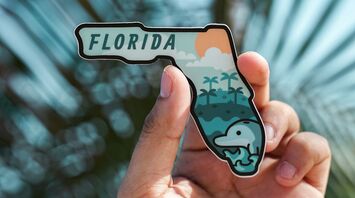 A person holding a colorful Florida-shaped souvenir magnet featuring a dolphin and palm trees