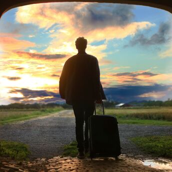 Man with suitcase walking towards a vibrant sunset through an arched tunnel