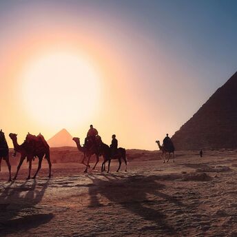 Four persons riding camels walking on sand beside pyramid