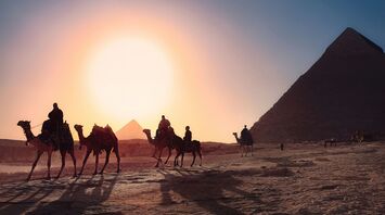 Four persons riding camels walking on sand beside pyramid