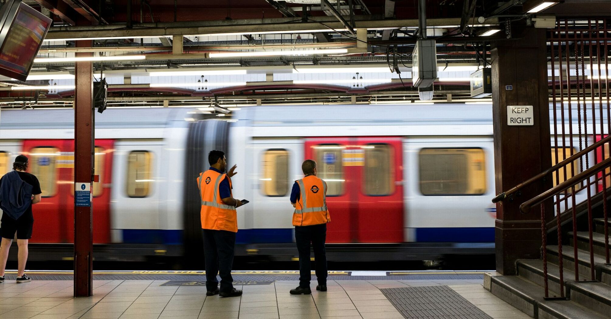 Two station workers in orange vests monitoring a rapidly moving train at an underground platform