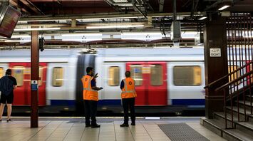 Two station workers in orange vests monitoring a rapidly moving train at an underground platform