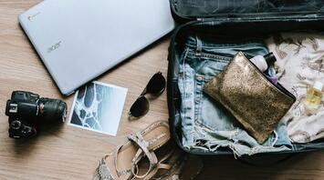 A suitcase partially packed with clothes, a laptop, a camera, sunglasses, and sandals