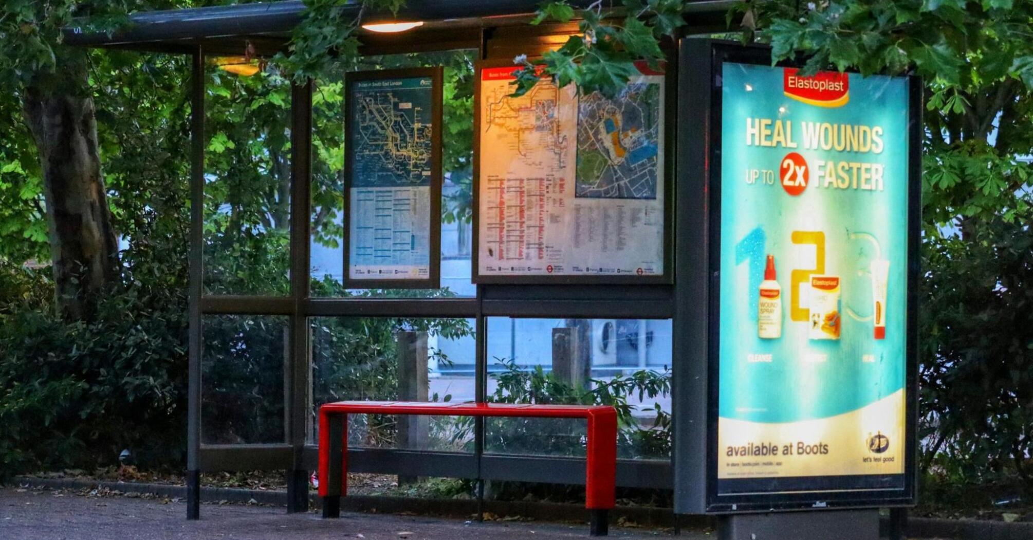 A well-lit bus stop with route maps and an advertisement poster, surrounded by green trees