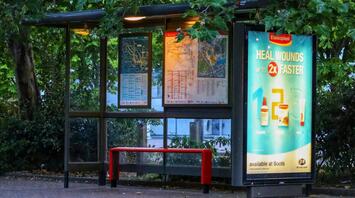 A well-lit bus stop with route maps and an advertisement poster, surrounded by green trees