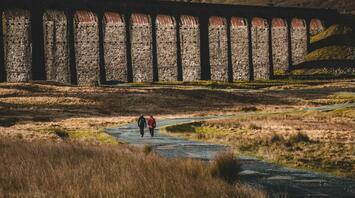 Two cyclists riding on a path beside a historic stone viaduct in the Yorkshire Dales