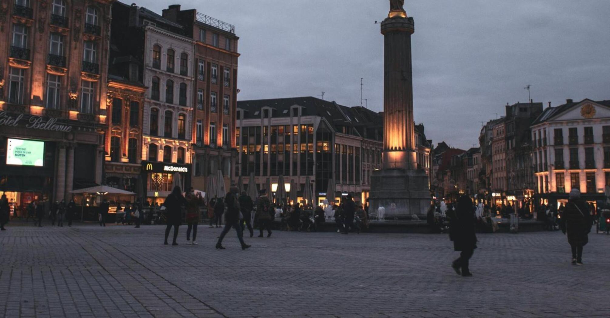 Evening view of a busy square in a French city with a tall statue and illuminated buildings
