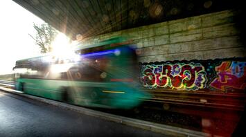 A green bus passes under a bridge with graffiti on the wall, sunlight shining through