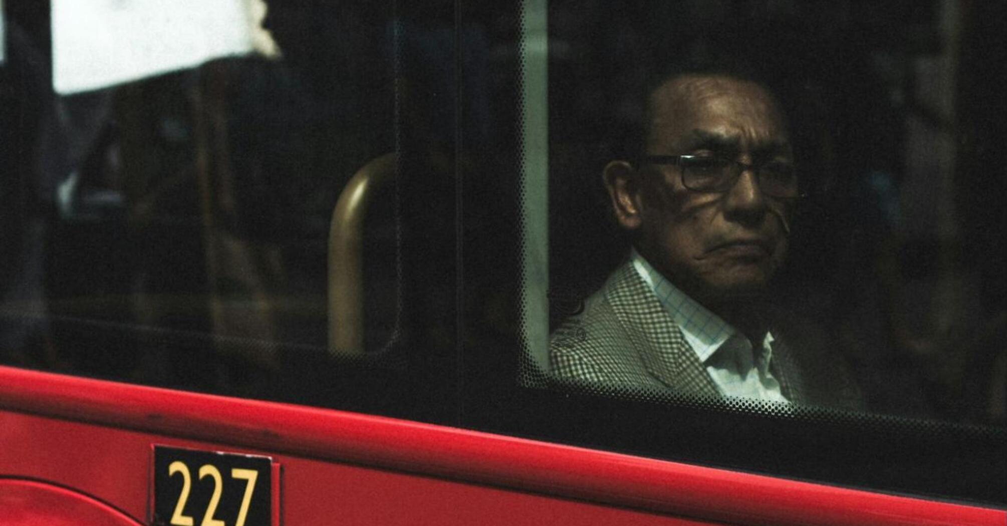 An older man sitting inside a red bus, looking out of the window