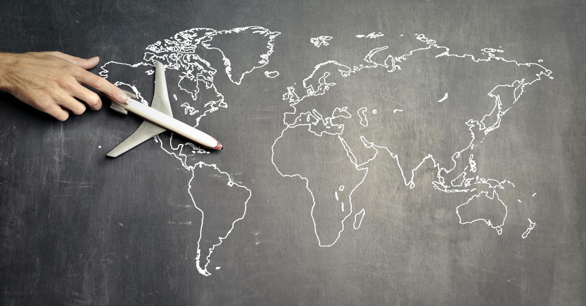 A hand guiding a toy airplane over a world map drawn on a chalkboard