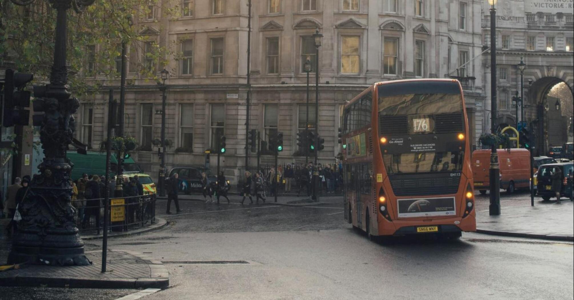 A double-decker bus travels through a busy city intersection in England, with historic buildings in the background and pedestrians crossing the street