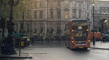 A double-decker bus travels through a busy city intersection in England, with historic buildings in the background and pedestrians crossing the street