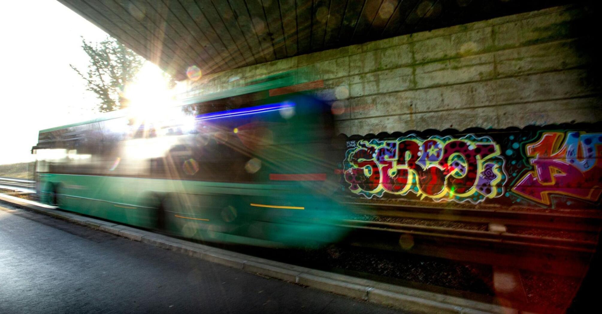 A green Stagecoach bus passing under a bridge with graffiti on the wall, sunlight flaring in the background