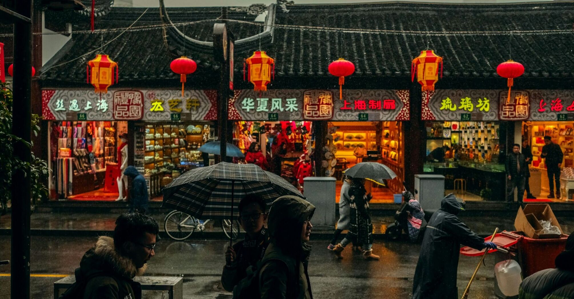 Rainy street scene in China with people holding umbrellas in front of traditional shops decorated with red lanterns