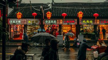 Rainy street scene in China with people holding umbrellas in front of traditional shops decorated with red lanterns