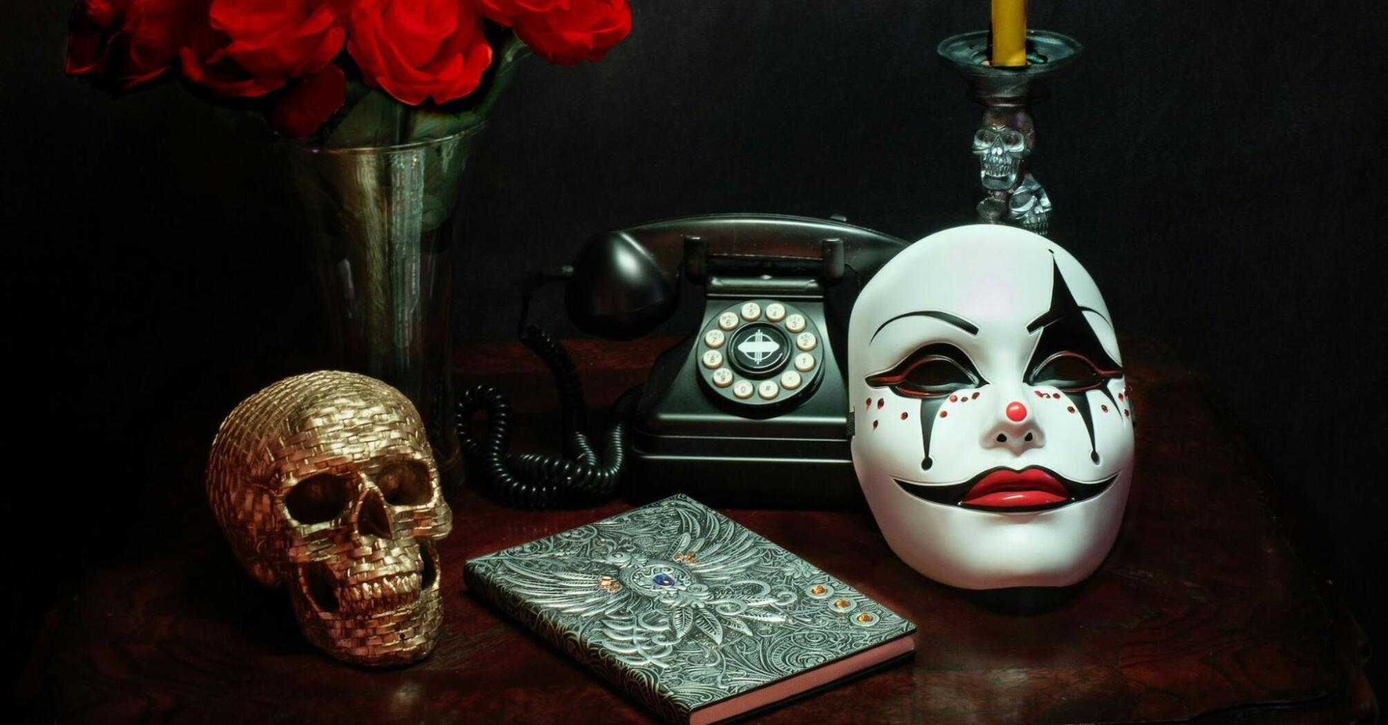 A vintage rotary phone, a theatrical mask, a golden skull, a bouquet of red roses, a candle, and an ornate journal arranged on a dark wooden table 