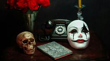 A vintage rotary phone, a theatrical mask, a golden skull, a bouquet of red roses, a candle, and an ornate journal arranged on a dark wooden table