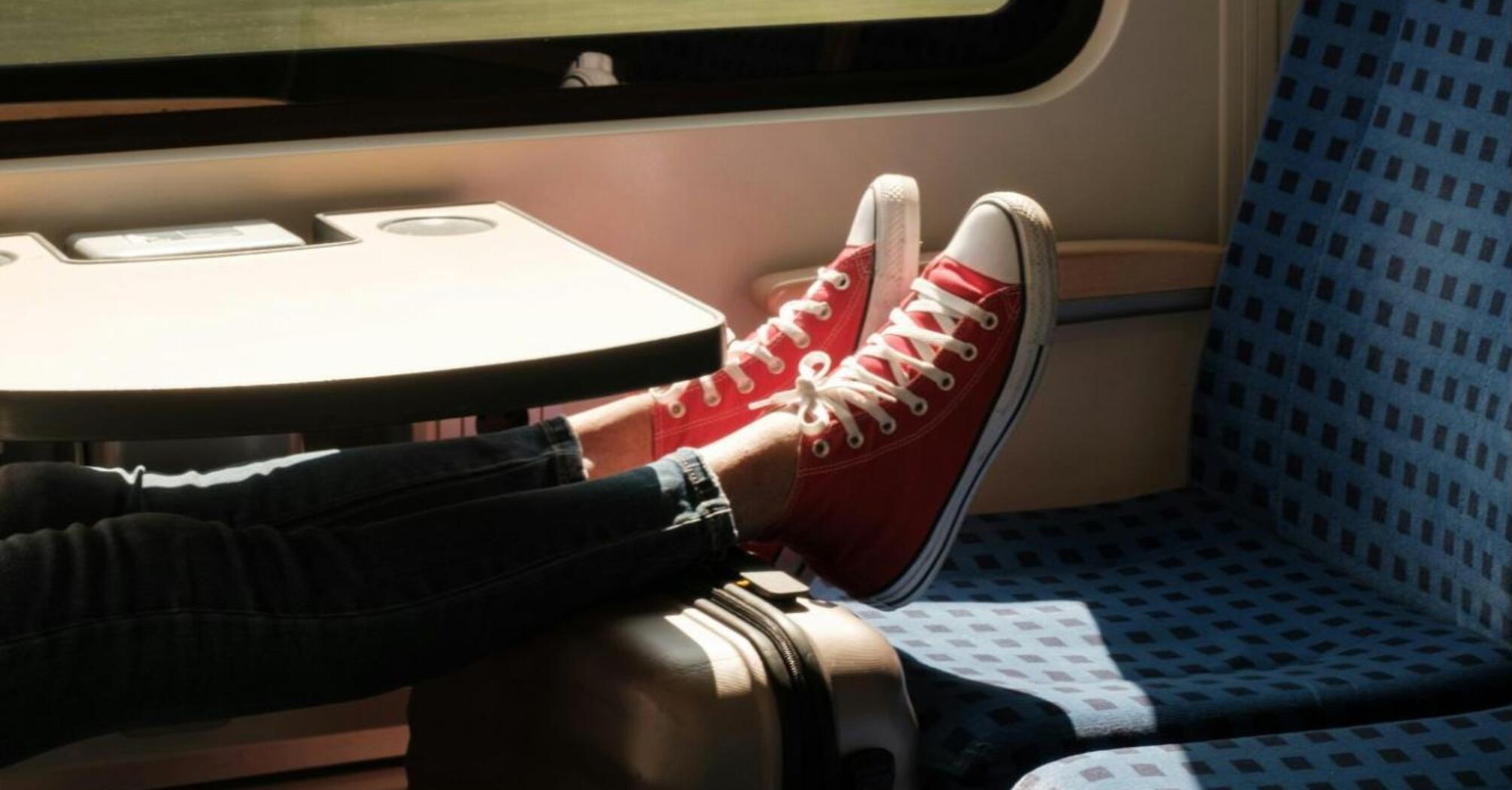 A person wearing red sneakers relaxes on a train, with their feet propped up on a suitcase next to a window showing blurred scenery
