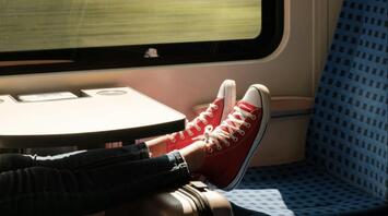 A person wearing red sneakers relaxes on a train, with their feet propped up on a suitcase next to a window showing blurred scenery