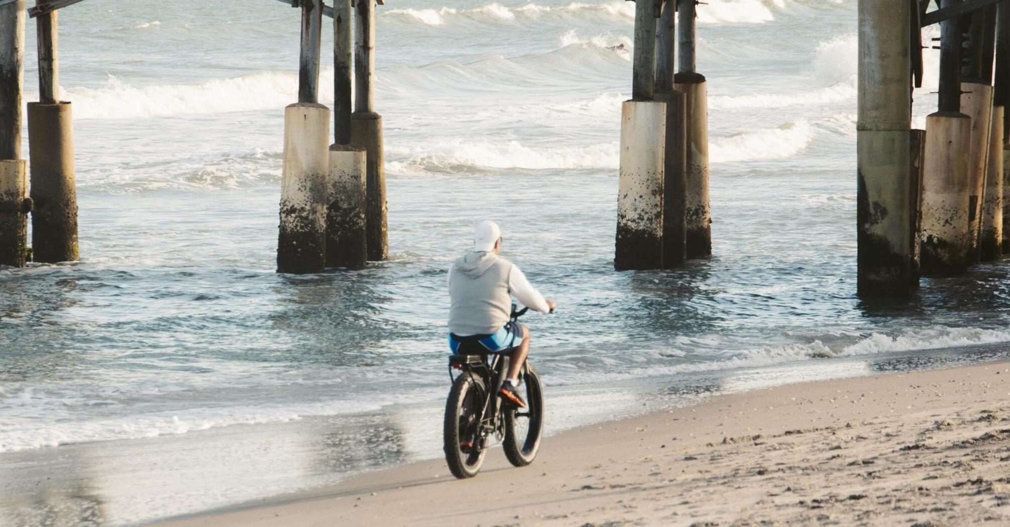 A person on a bicycle rides along the beach under a wooden pier