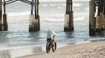 A person on a bicycle rides along the beach under a wooden pier