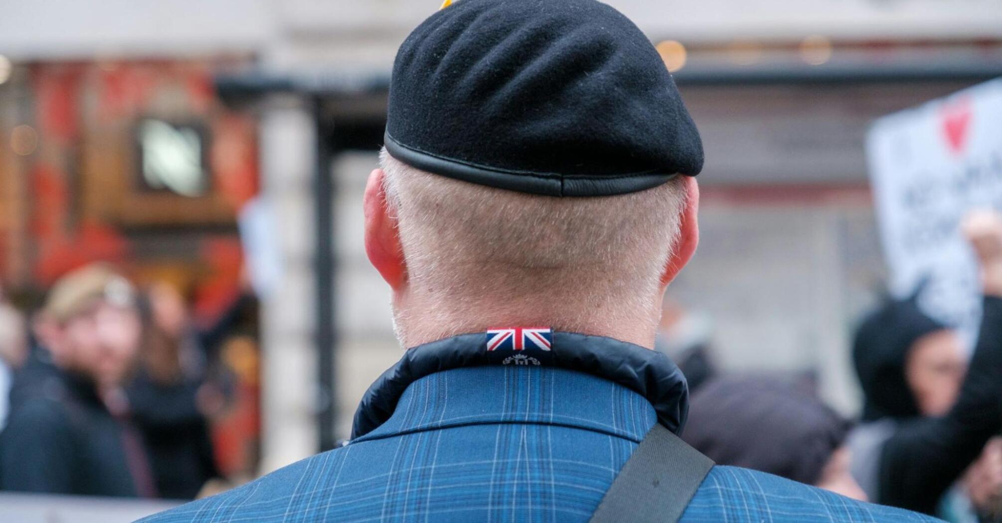 A veteran in a beret with a Union Jack emblem on his jacket collar, seen from behind at a public event