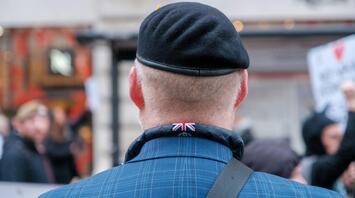 A veteran in a beret with a Union Jack emblem on his jacket collar, seen from behind at a public event