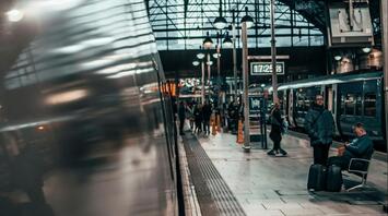 Passengers at a busy train station platform, with a train on the left and a high, arched glass ceiling above