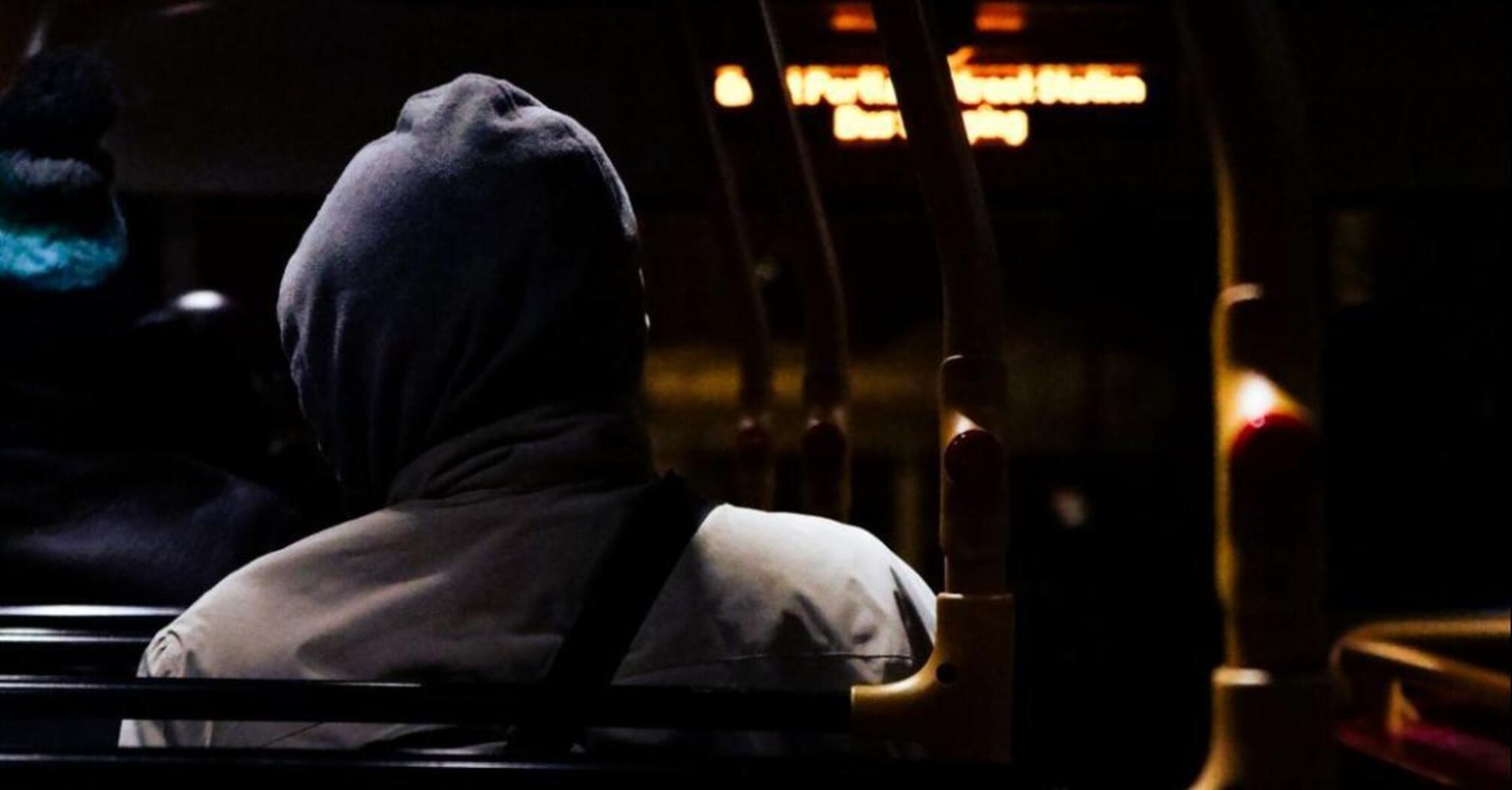 A passenger in a hoodie sits on a dimly lit bus at night