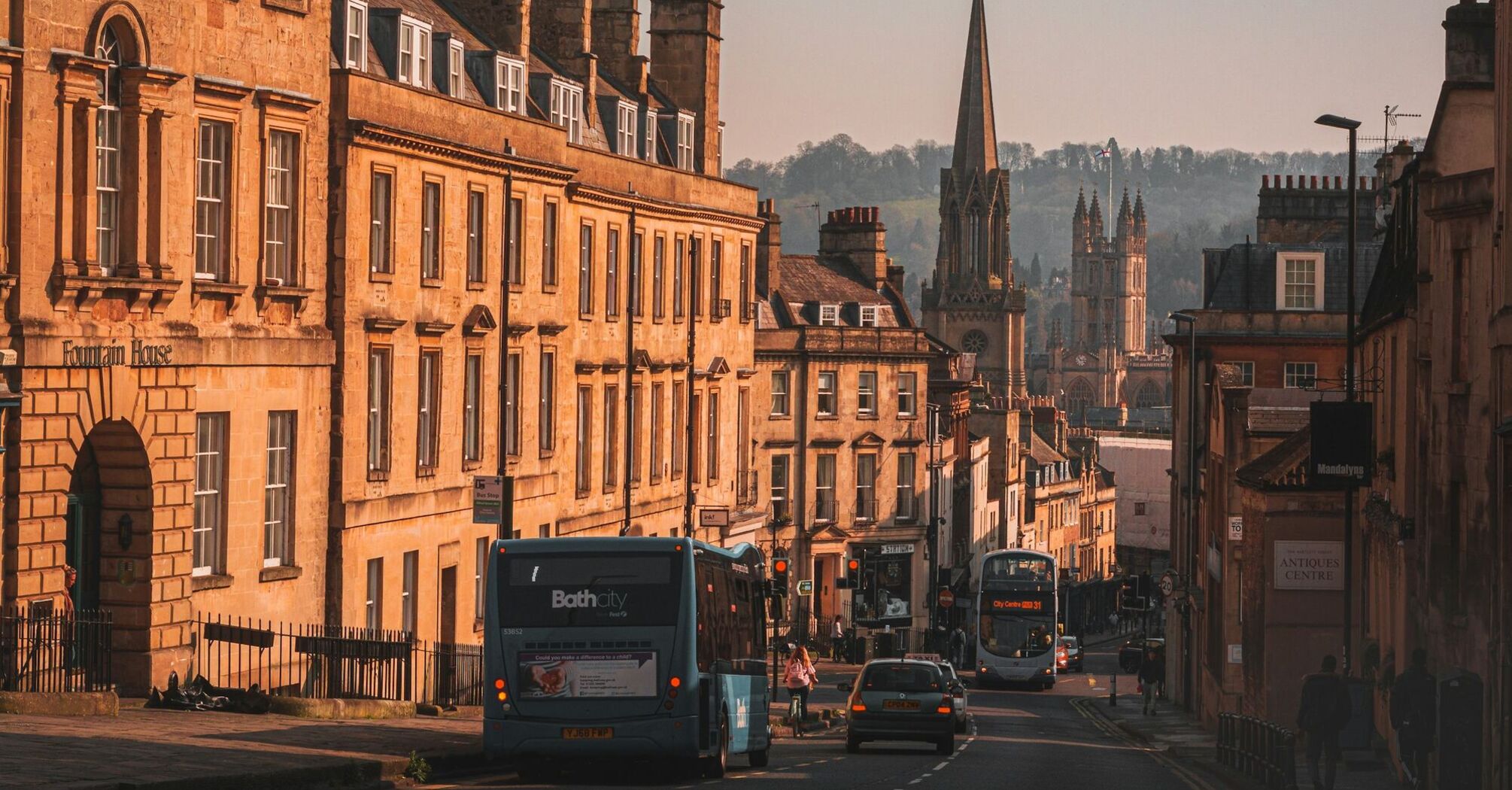 View of a historic street in the South West, showing buses, cars, and old buildings with a church in the background