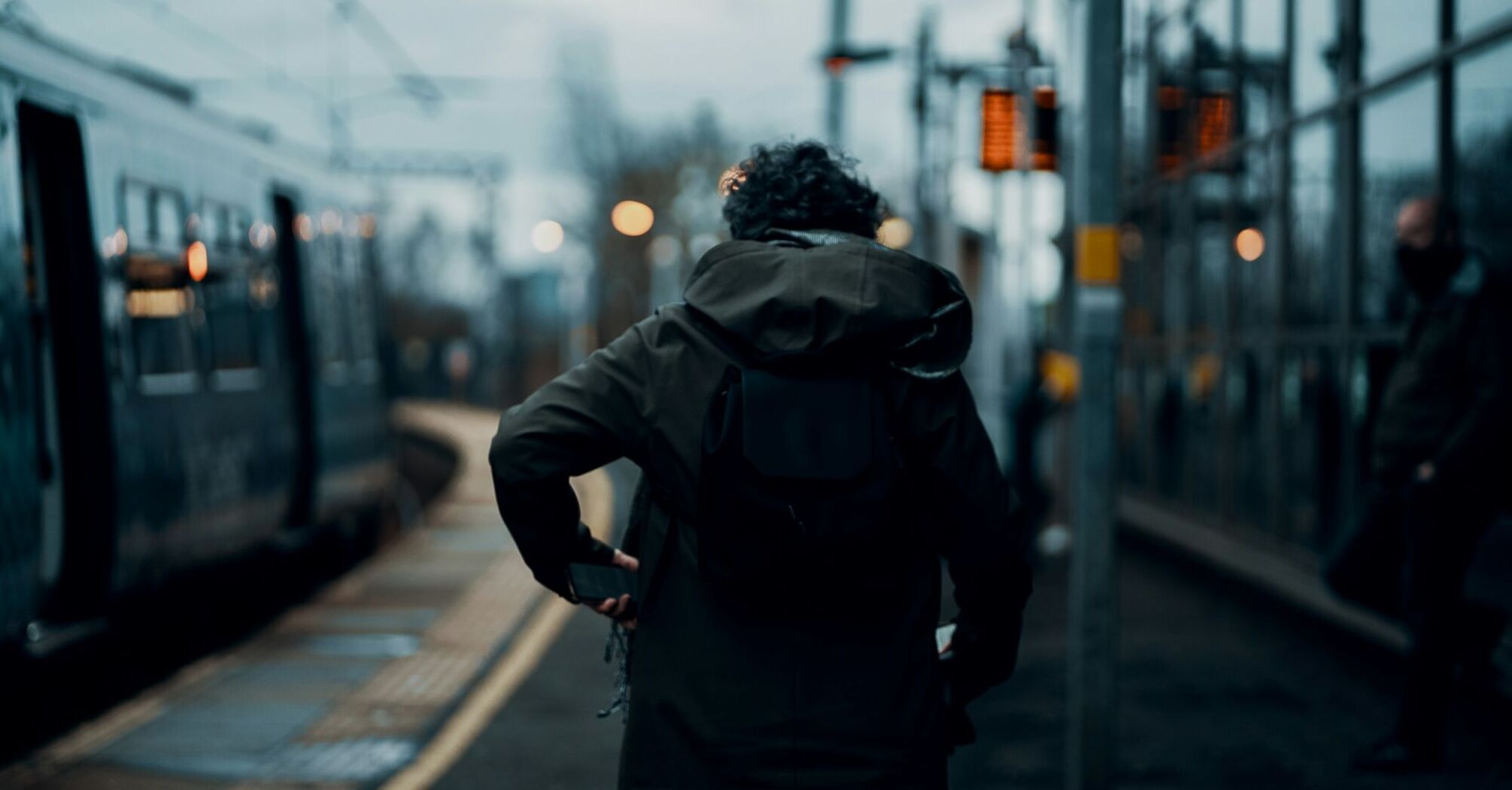 A person with a backpack waits on a train platform as a train arrives in the early morning or late evening