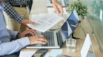 Two men working on a laptop with printed documents in an office